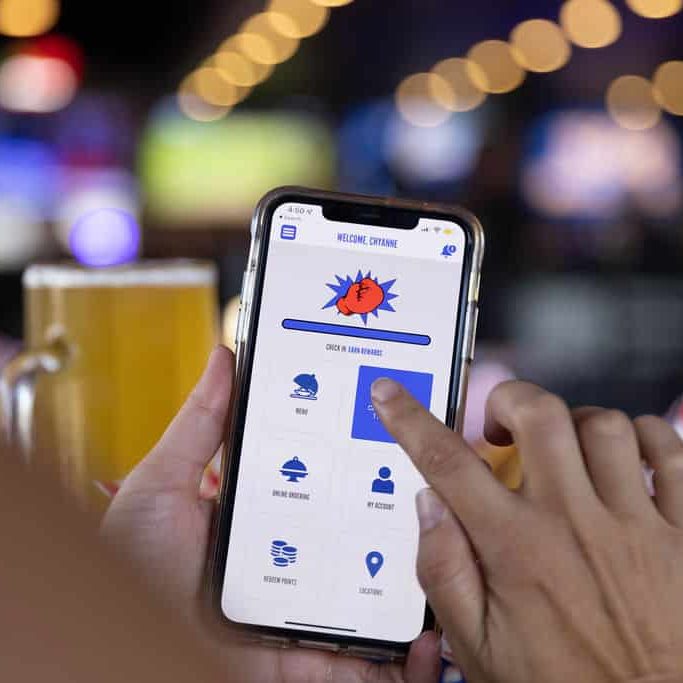 Hands holding cellphone using app, finger touching button on screen with beer and bar lights in the background.