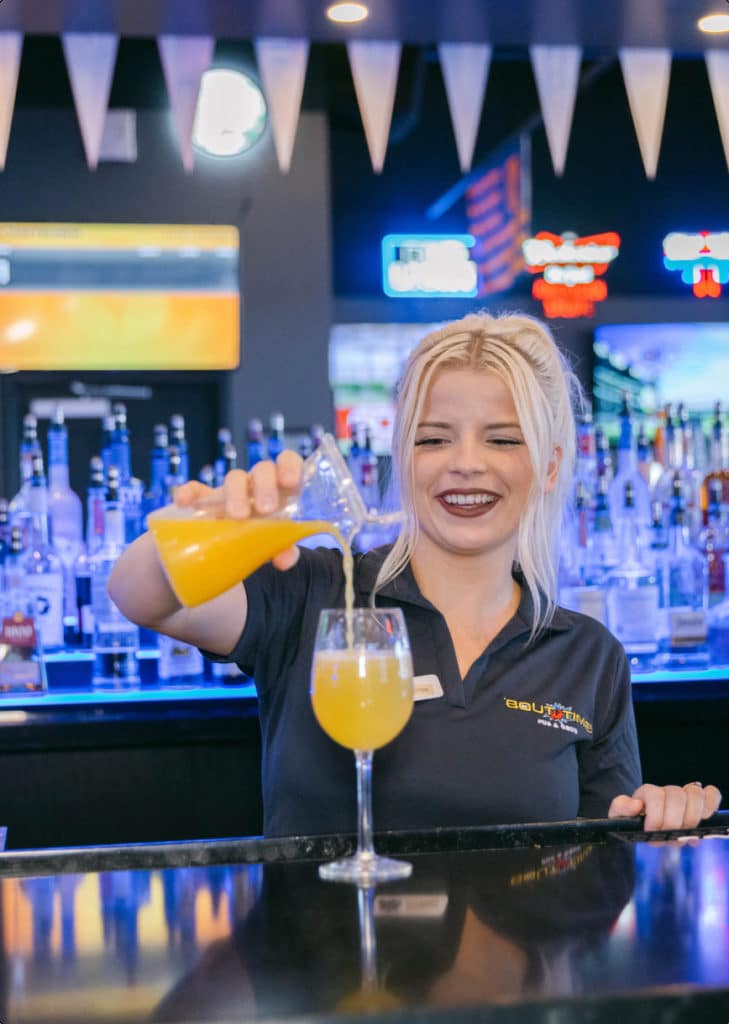 Bout Time Pub & Grub female employee pouring mimosa at bar while smiling.