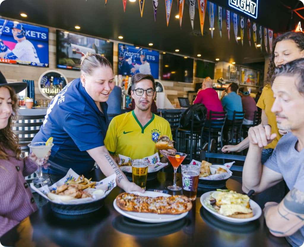 Customers at pub sitting at table, being served food by friendly waiter. Sports team flags and TVs hanging on the walls in the background.