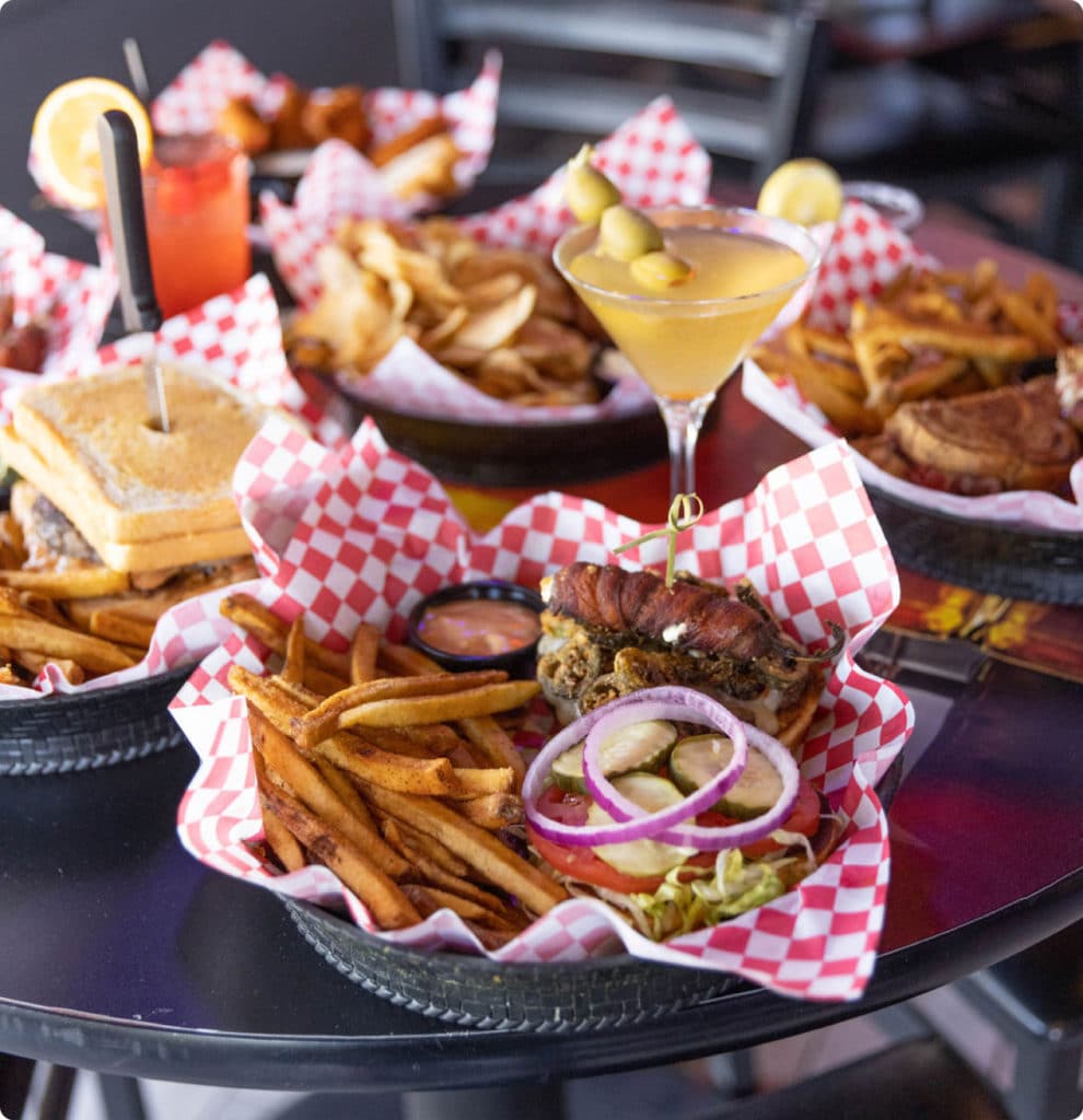 Table full of pub food. Food in baskets including open-face burger, fries, sandwich, martini.