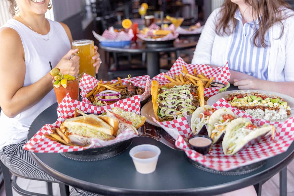 Pub table with two customers sitting at it, one holding a beer. large baskets of food on the table including sandwich, fries, burger, tacos.